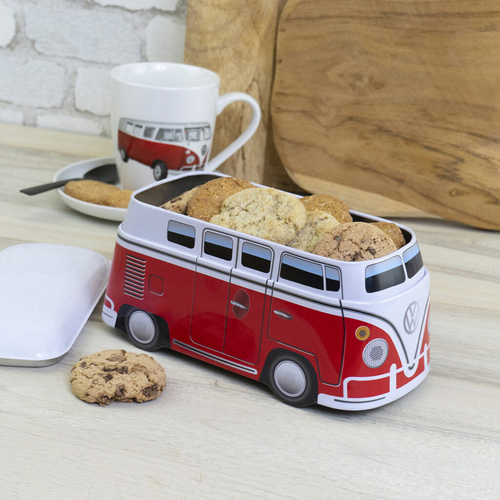 Eine rot-weiße Keksdose in Form eines alten Lieferwagens ist mit Keksen gefüllt. Ein Schokoladenkeks liegt daneben. Im Hintergrund steht eine Tasse mit einem passenden Van-Motiv auf einer weißen Untertasse. Ein Holzbrett ist ebenfalls zu sehen.