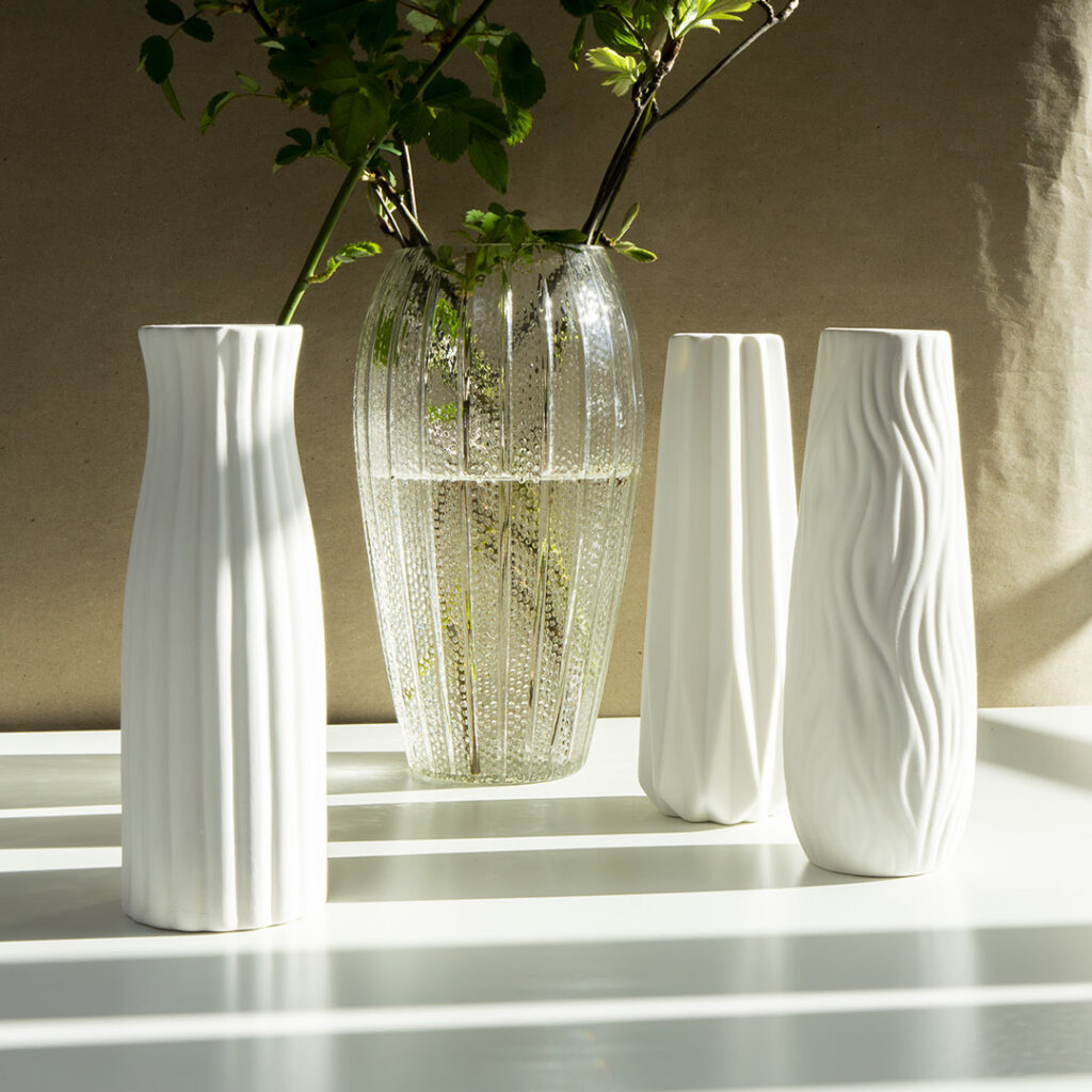 Vier Vasen auf einer weißen Fläche: drei sind hoch, weiß und aus Keramik mit unterschiedlicher Struktur, und eine ist eine Vase aus klarem Glas, die Wasser und grüne Blätterzweige enthält. Das Sonnenlicht wirft weiche Schatten auf die Szene.