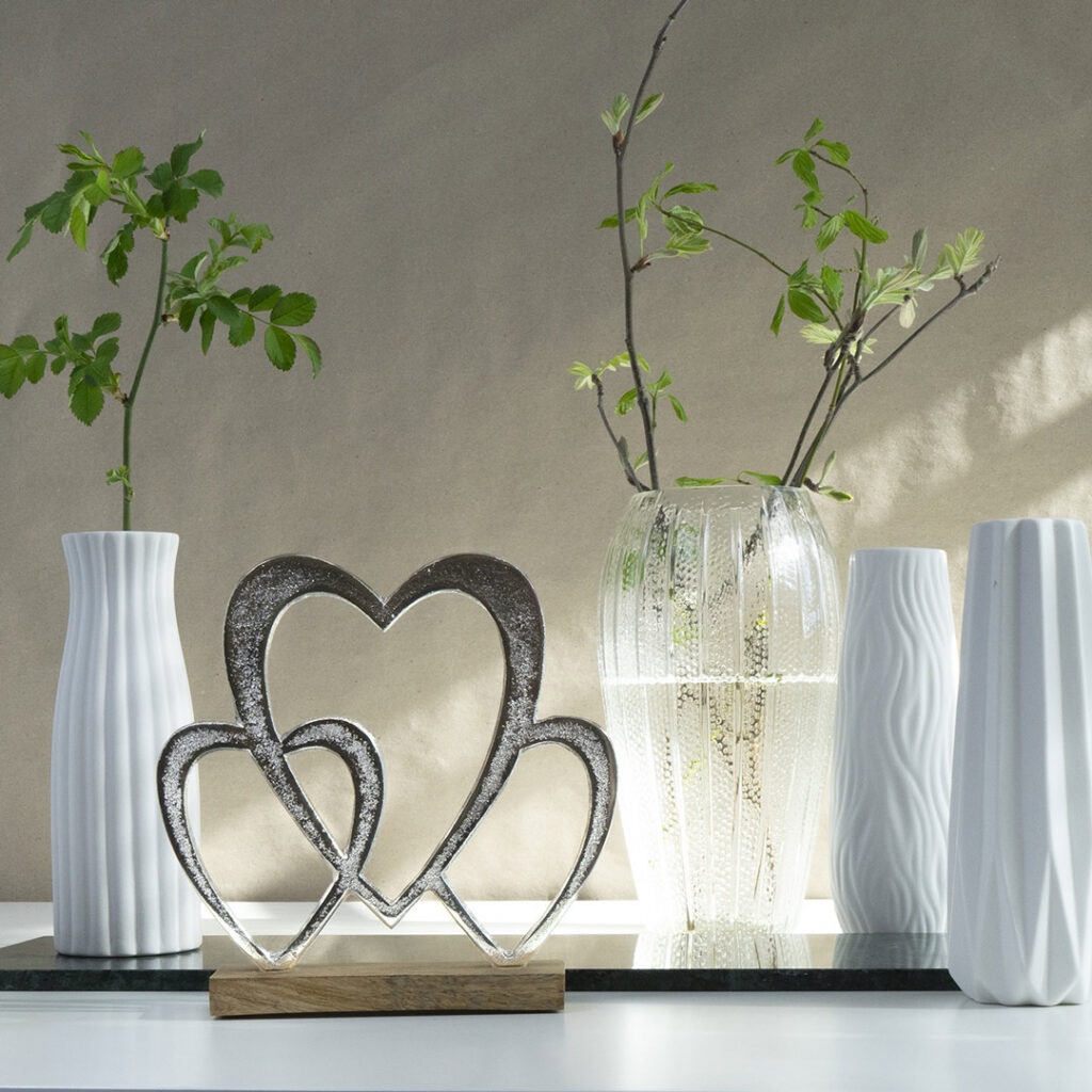 Ein dekoratives Arrangement mit drei weißen Vasen und einer Klarglasvase mit grünen Blätterzweigen sowie einer Metallskulptur mit drei ineinander verschlungenen Herzen auf einem Holzsockel, alles auf einer weißen Fläche.
