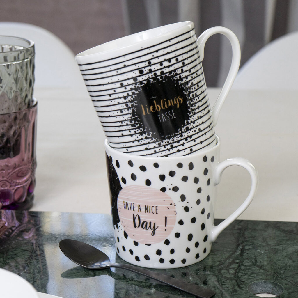 Zwei gestapelte Keramikbecher stehen auf einem Tisch. Der obere Becher hat schwarze Querstreifen und goldene Schrift, der untere Becher ist weiß mit schwarzen Punkten und der Aufschrift "Have a nice day!" in Rosa. Ein Löffel und ein strukturiertes Glas stehen daneben.