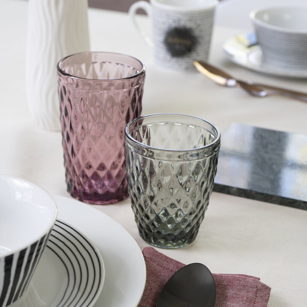 Zwei strukturierte Trinkgläser, ein lila und ein graues, stehen auf einem gedeckten Esstisch mit gestreiften Schüsseln, Tellern, einer Tasse, einem Löffel und einer rosa Serviette auf einer weißen Tischdecke.