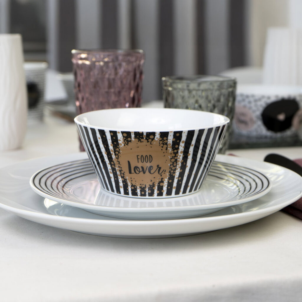 Eine Keramikschale mit schwarzen und weißen Längsstreifen und dem Schriftzug Food Lover in der Mitte steht auf einem passenden Teller und einem großen weißen Teller, umgeben von strukturierten Glastassen auf einem gedeckten Esstisch.