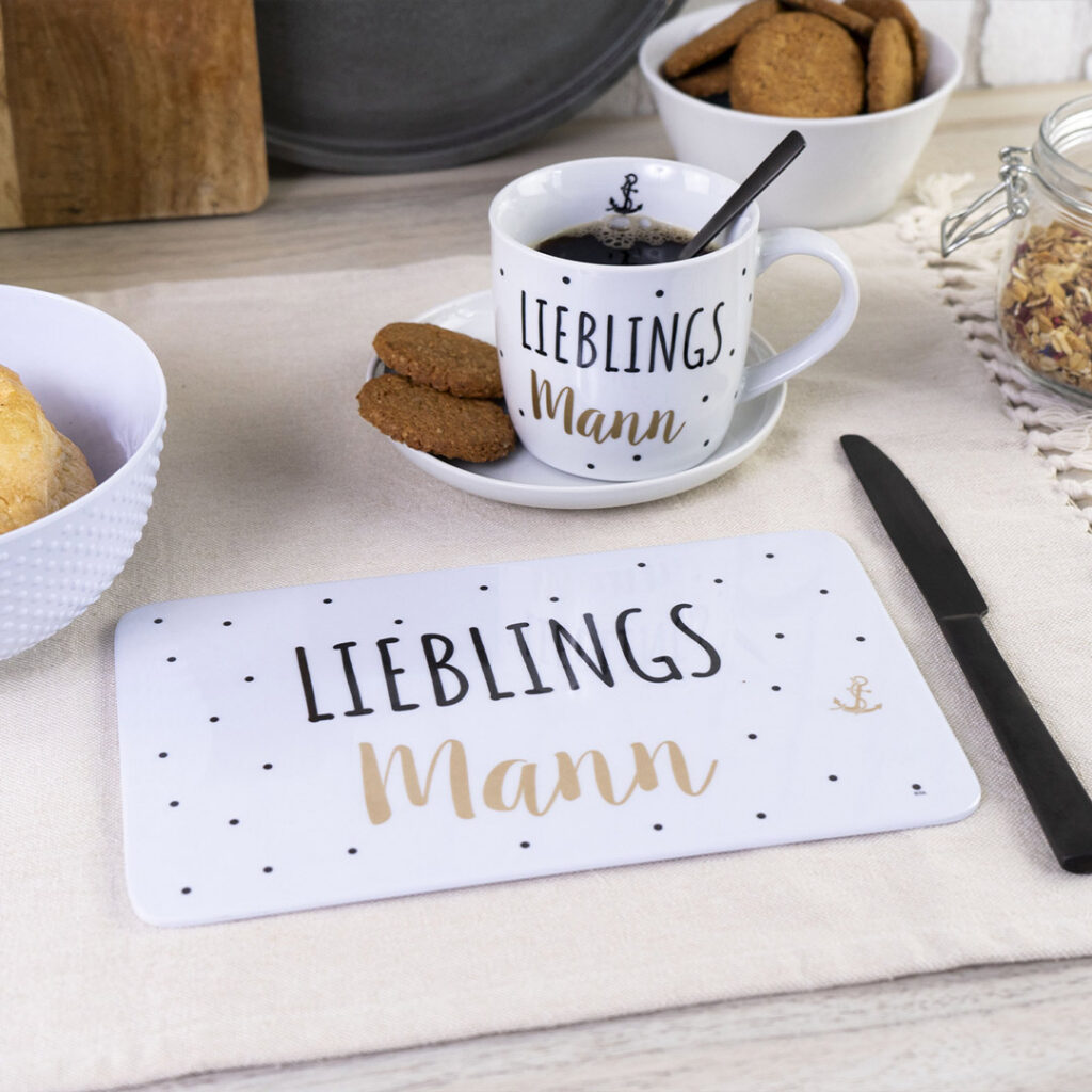 Ein gedeckter Frühstückstisch mit einer Kaffeetasse, einer Untertasse und einem Tischset, auf denen auf Deutsch "Lieblingsmann" steht. Auf einem hellen Tischset liegen Kekse, eine Schüssel mit Müsli, ein Brötchen und ein Messer.