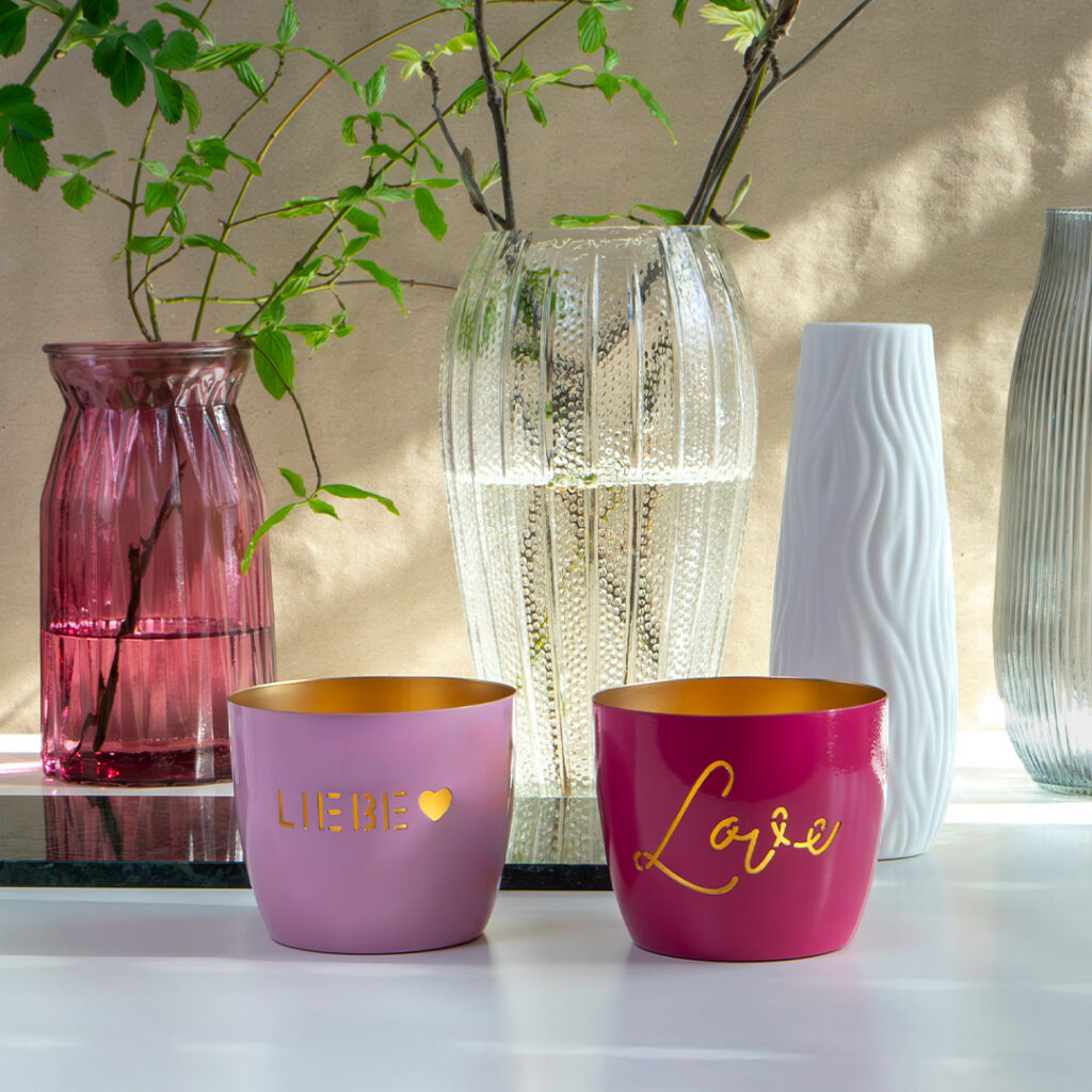 Zwei dekorative Tassen, eine hellrosa mit der Aufschrift LIEBE und einem Herz, die andere dunkelrosa mit der Aufschrift Liebe in Gold, stehen auf einem Tisch vor Glas- und Keramikvasen mit grünen Zweigen.