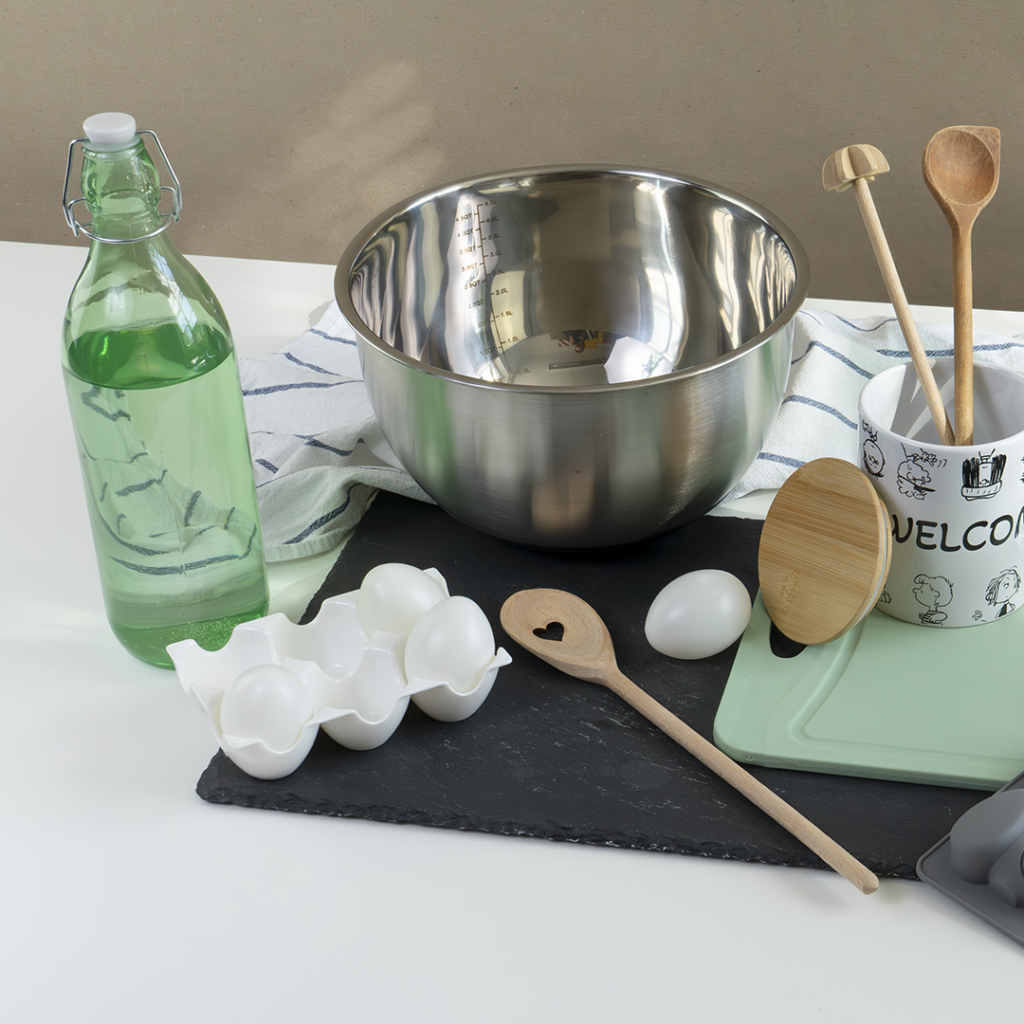 Eine Küchenszene mit einer Flasche grüner Flüssigkeit, vier Eiern in einem Karton, einer Rührschüssel, Holzlöffeln, einem Becher, einem Bambusdeckel, einem Schneidebrett und einem gestreiften Handtuch auf einem weißen Tisch.