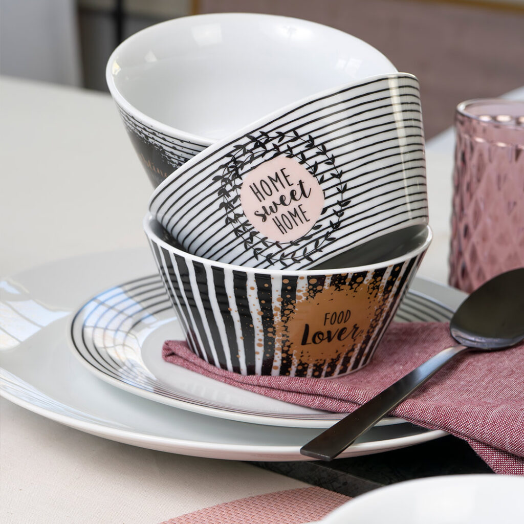 Zwei gemusterte Schalen, die auf weißen Tellern stehen, wobei eine Schale die Aufschrift HOME sweet HOME und die andere FOOD lover trägt. Auf dem Tisch liegen eine rosa Serviette, ein Löffel und ein strukturiertes rosa Glas.
