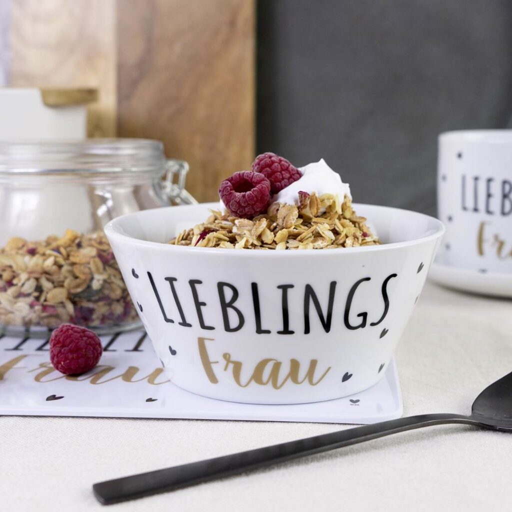 Eine weiße Schale mit der Aufschrift Lieblings Frau, gefüllt mit Müsli, Himbeeren und Joghurt, steht auf einem Tisch neben einem schwarzen Löffel, mit einem Glas Müsli und einem Becher im Hintergrund.