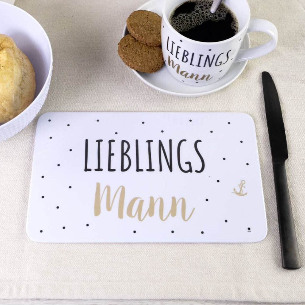 Frühstückstisch mit einem Teller mit Brot, einer Tasse Kaffee, Keksen, einem Messer und einem Tischset. Auf der Tasse und dem Untersetzer steht in Schwarz und Gold der Schriftzug Lieblingsmann.