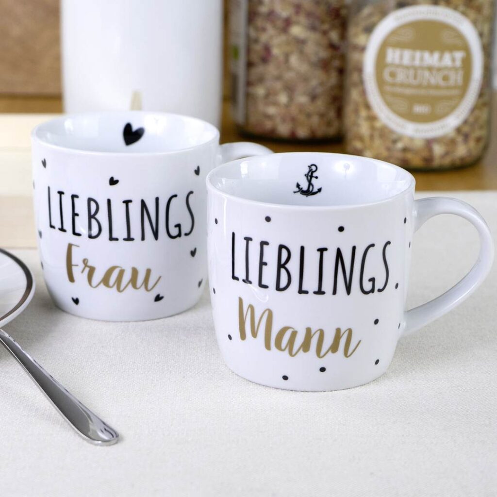 Zwei weiße Tassen auf einem Tisch; auf einer steht LIEBLINGS Frau mit kleinen Herzen, auf der anderen LIEBLINGS Mann mit einem Anker und Punkten. Im Hintergrund stehen Müslidosen und ein Löffel.