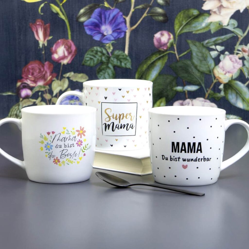 Drei weiße Tassen mit deutschen Botschaften für Mütter sind auf einem Tisch mit einem Löffel angeordnet. Als Hintergrund dient eine Blumentapete. Die Tassen zeigen dekorative Muster und positive Sprüche wie Super Mama und Du bist wunderbar.