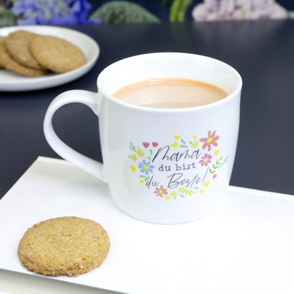 Ein weißer Becher mit floralen Motiven und Text steht auf einem Buch neben einem Keks. Der Becher ist mit einem hellbraunen Getränk gefüllt. Im Hintergrund befindet sich ein Teller mit mehreren Keksen.