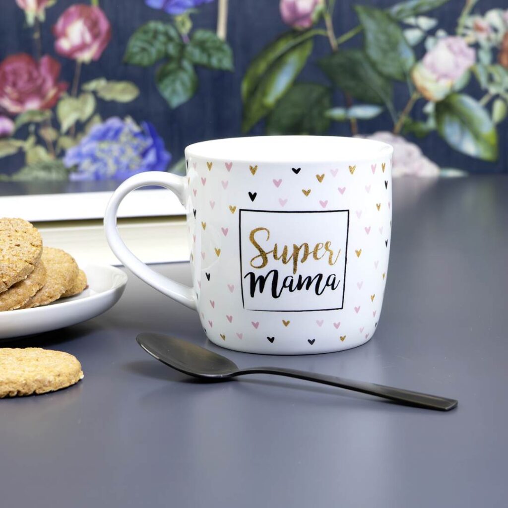 Eine weiße Tasse mit kleinen Herzmustern und der Aufschrift Super Mama in Gold und Schwarz steht neben einem schwarzen Löffel und einem Teller mit Keksen auf einem grauen Tisch mit einem Blumenbild im Hintergrund.