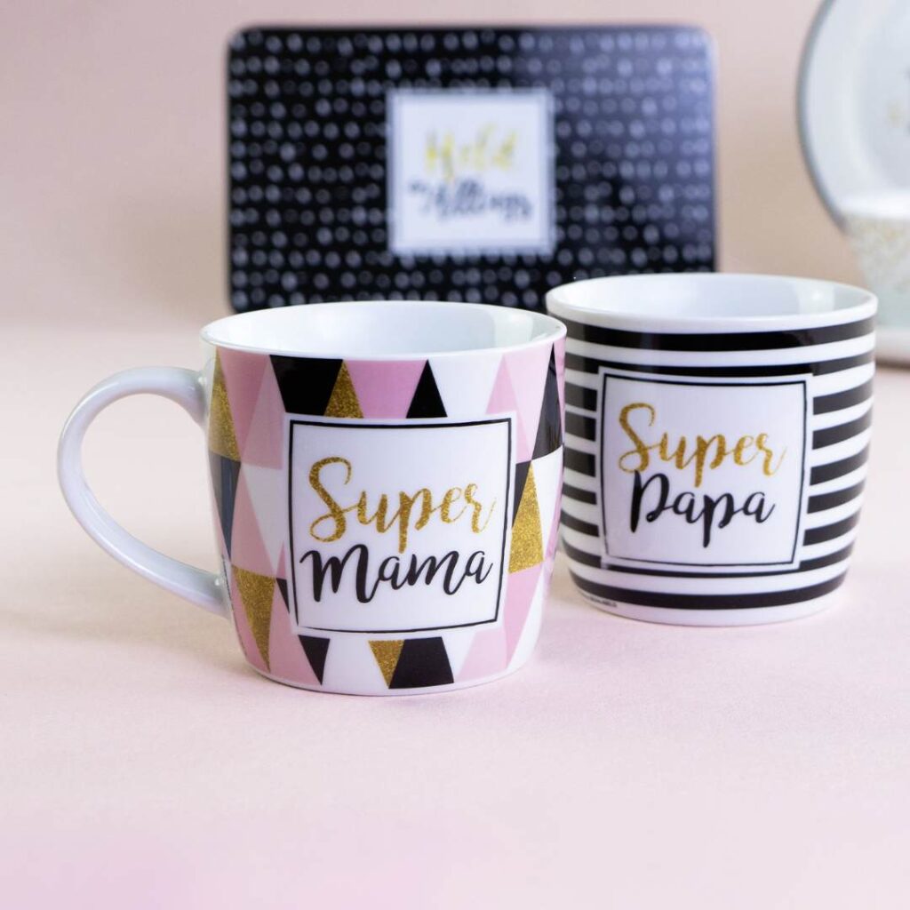 Zwei Keramiktassen auf einer rosa Fläche: eine mit rosa, weißen, schwarzen und goldenen Dreiecken mit der Aufschrift Super Mama und eine mit schwarzen und weißen Streifen mit der Aufschrift Super Papa. Im Hintergrund ist eine gepunktete Schachtel zu sehen.