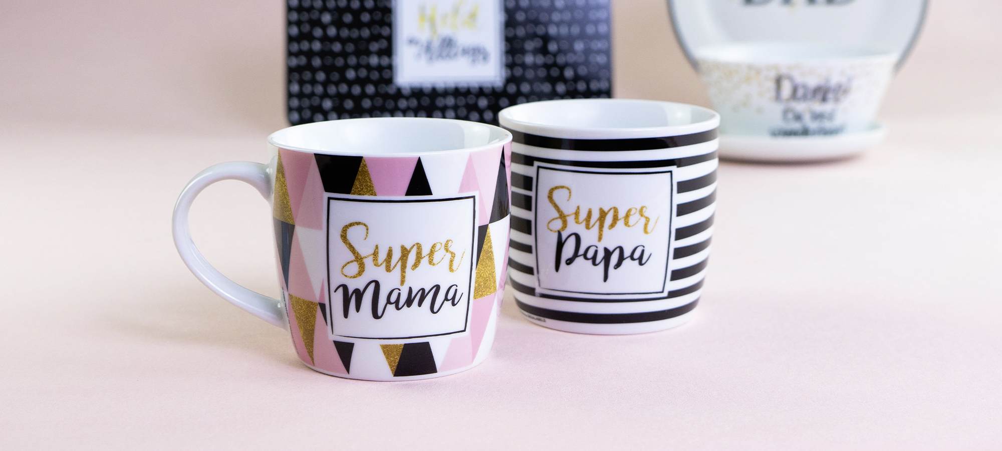 Zwei Tassen auf einer hellrosa Fläche; eine hat rosa, schwarze und goldene Dreiecke und steht Super Mama, die andere hat schwarze und weiße Streifen und steht Super Papa. Im Hintergrund sind unscharfe Tassen zu sehen.