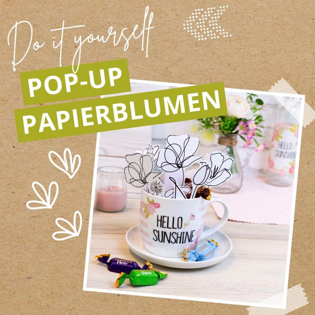 Ein DIY-Projekt mit einem weißen Becher mit dem Text "HELLO SUNSHINE", der Papierblumen hält, auf einer Untertasse mit Süßigkeiten. Im Hintergrund sind Blumen in einer Vase und eine rosa Kerze zu sehen. Der Text lautet: Do it yourself POP-UP PAPIERBLUMEN.