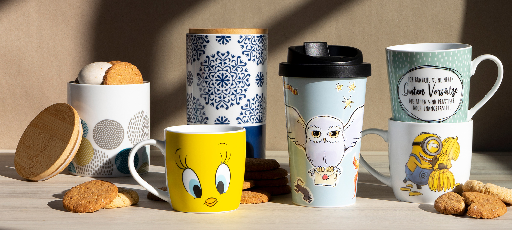 Fünf Tassen und Becher mit Keksen, die mit Cartoon-Motiven und Mustern versehen sind. Zu den Motiven gehören Tweety Bird, Hedwig die Eule, Minions und blaue geometrische Muster. Die Kekse sind um die Tassen herum auf einer hellen Oberfläche verstreut.
