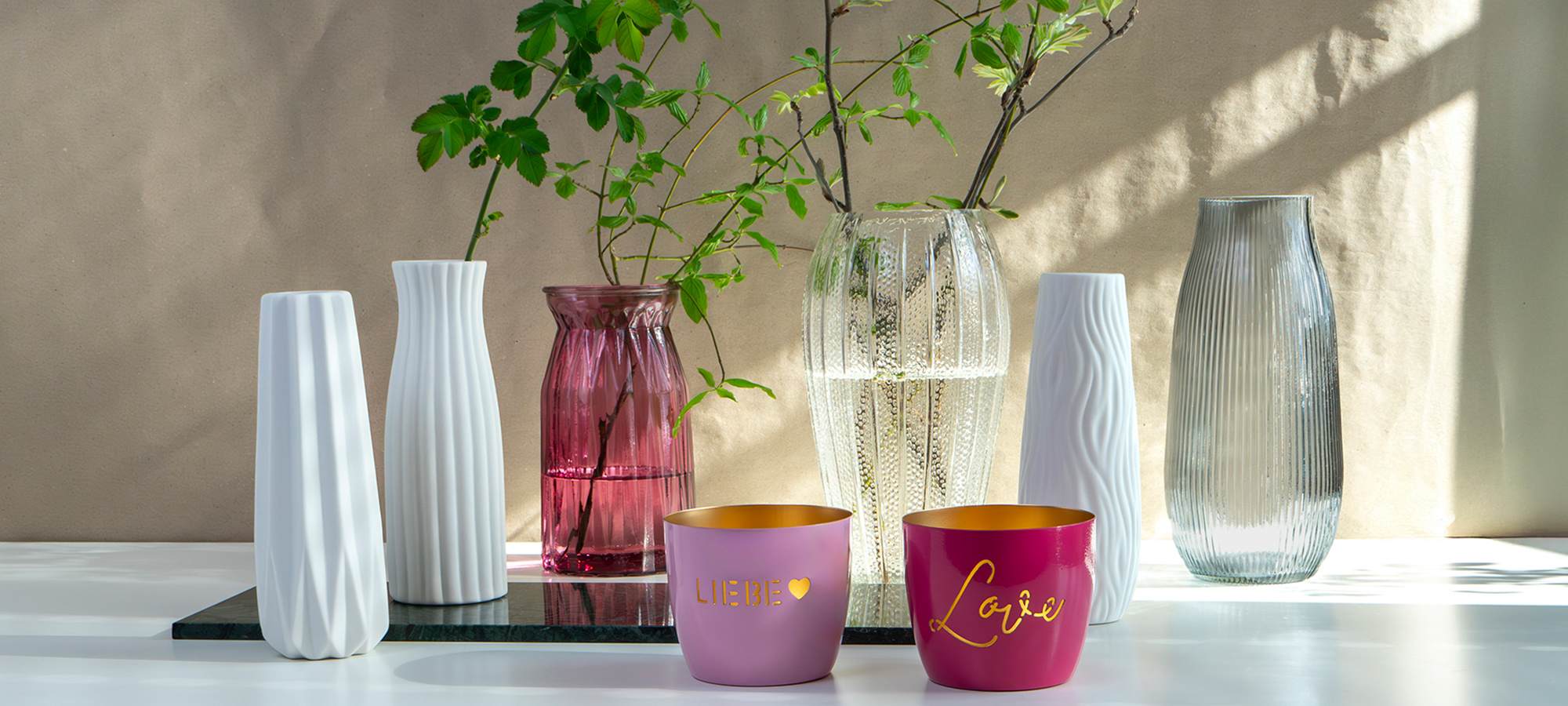Auf einer weißen Fläche steht eine Gruppe strukturierter Glas- und Keramikvasen, einige mit blattartigen Zweigen. Davor stehen zwei rosafarbene Kerzenhalter mit der Aufschrift LIEBE und Liebe. Das Sonnenlicht wirft Schatten auf die Szene.