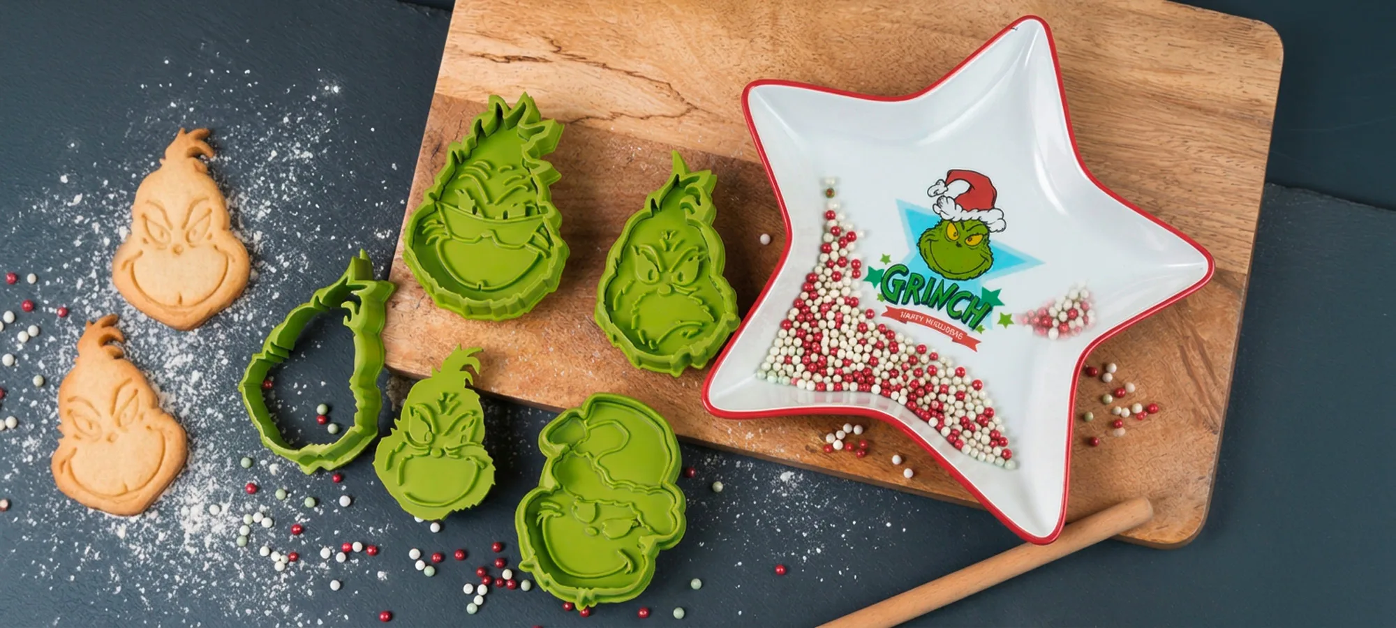 Zwei Grinch-förmige Zuckerkekse, mehrere grüne Grinch-Ausstechformen und eine sternförmige Schale, gefüllt mit roten, weißen und grünen Streuseln, auf einem Holzschneidebrett.