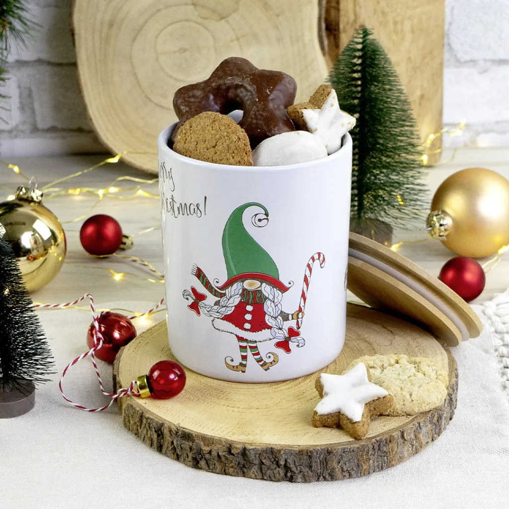 Weiße Tasse mit der Abbildung eines Weihnachtswichtels, gefüllt mit verschiedenen Weihnachtsplätzchen und umgeben von Weihnachtsschmuck, Mini-Weihnachtsbäumchen und einer Holzscheibe.