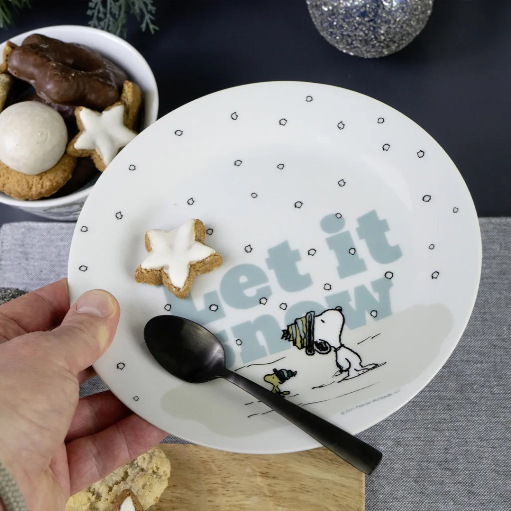 Eine Hand hält einen Teller mit dem Snoopy-Motiv Let it snow, auf dem ein schwarzer Löffel, ein sternförmiger Keks und im Hintergrund eine Schale mit verschiedenen Keksen zu sehen sind.