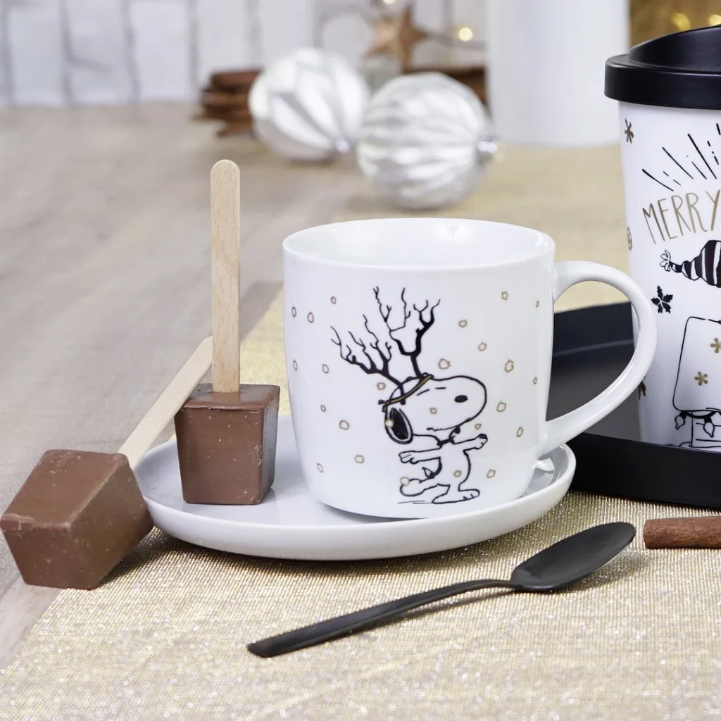Eine Snoopy-Tasse mit passender Untertasse, zwei Rührstäbchen für heiße Schokolade und einem schwarzen Löffel auf einem Tisch.