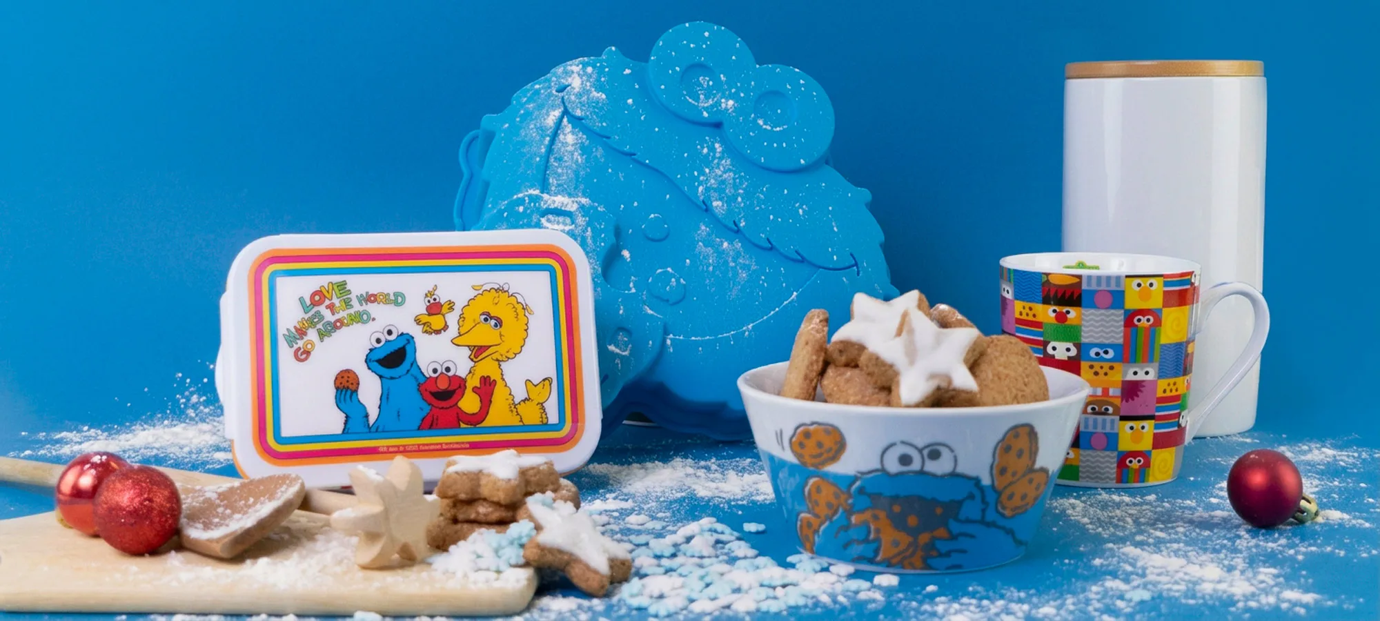 Eine Snackbox im Sesamstraße-Design, eine Schale mit sternförmigen Keksen und eine Tasse, dazu eine blaue Silikonform in Form des Krümelmonsters auf einer blauen, mit Mehl bestäubten Fläche.