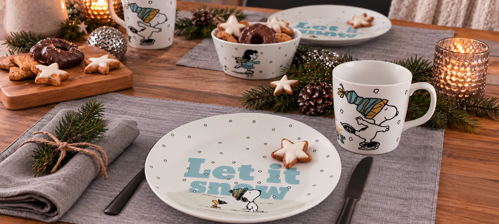Tassen und Teller mit Peanuts-Motiven, verziert mit Snoopy und dem Schriftzug Let it snow, angeordnet auf einem Tisch mit Weihnachtsplätzchen und Tannenzweigen.