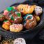Ein Teller mit Halloween-Keksen in Form von Totenköpfen und Gesichtern, verziert mit buntem Zuckerguss und Streuseln, steht auf einer dunklen Fläche mit weiteren Totenkopfkeksen in der Nähe.