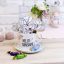 Ein weißer Becher mit der Aufschrift HELLO SUNSHINE ist mit Blumen aus Papier beklebt. Die Tasse steht auf einer Untertasse mit eingewickelten Bonbons. In der Nähe stehen Kerzen, eine kleine Vase mit frischen Blumen und eine Flasche auf einem hellen Holztisch.