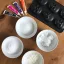 Vier Tuben Lebensmittelfarbe, eine Silikonform in Totenkopfform und vier Schüsseln mit Zucker, Kokosöl und anderen weißen Backzutaten auf einem Holztisch.