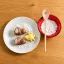 Zwei eichelförmige Pralinen und ein gelbes, sternförmiges Bonbon auf einem weißen Teller mit Puderzucker, daneben eine Schale mit Zucker und ein Holzlöffel mit einem herzförmigen Ausschnitt.